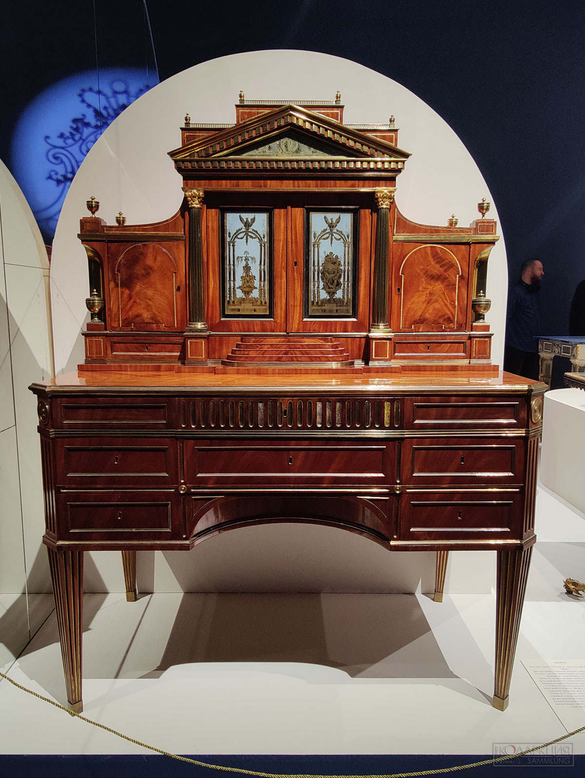 Бюро. Россия. Санкт-Петербург, мастерская Г. Гамбса. 1800-e. Дуб (основа), красное дерево, латунь, бронза, стекло, сафьян; фанеровка, штамповка, литьё, эгломизе, гравировка, тиснение. Бюро в «стиле жакоб» является эталонным для подобных русских краснодеревных бюро. Аналогичные предметы поставлял в Россию в 1780-х гг. мебельщик Д. Рентген, а Петербургский столяр Х. Мейер начал изготавливать похожие образцы для русского двора уже в 1790-е гг. Г. Гамбс, ученик Д. Рентгена, заимствовал у него многие конструктивные и художественные приёмы. Стеклянные вставки, выполненные в технике эгломизе, один из характерных приёмов декорирования корпусной мебели 1790-1800-х гг.